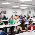 Classroom filled with students seated at tables participating in a clinical skills session, with training materials and supplies spread across the room.