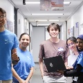 Group of students standing in a clinical classroom, one holding a laptop while others gather nearby, participating in a collaborative group activity.